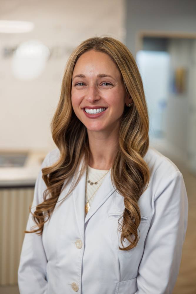 Portrait of Dr. Samantha Rawdin wearing a white coat and smiling in a dental clinic – Andrew Koenigsberg