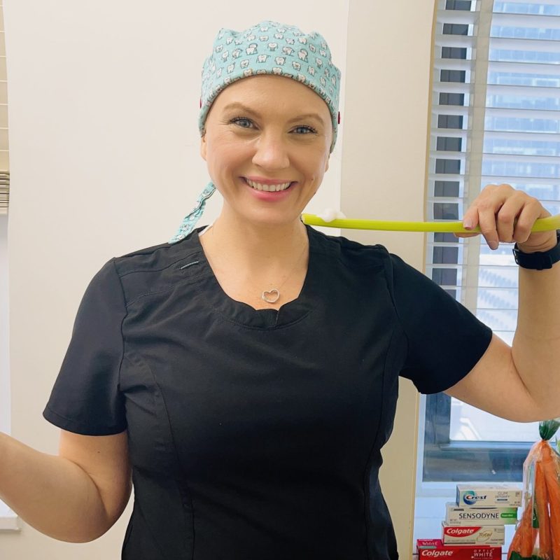 A smiling dental professional holding a long green toothbrush-like tool while standing in a dental office, representing tips on how to brush teeth without toothbrush – how to brush teeth without toothbrush